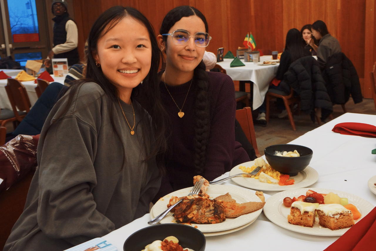 Two students sit at a table smiling, with plates of food