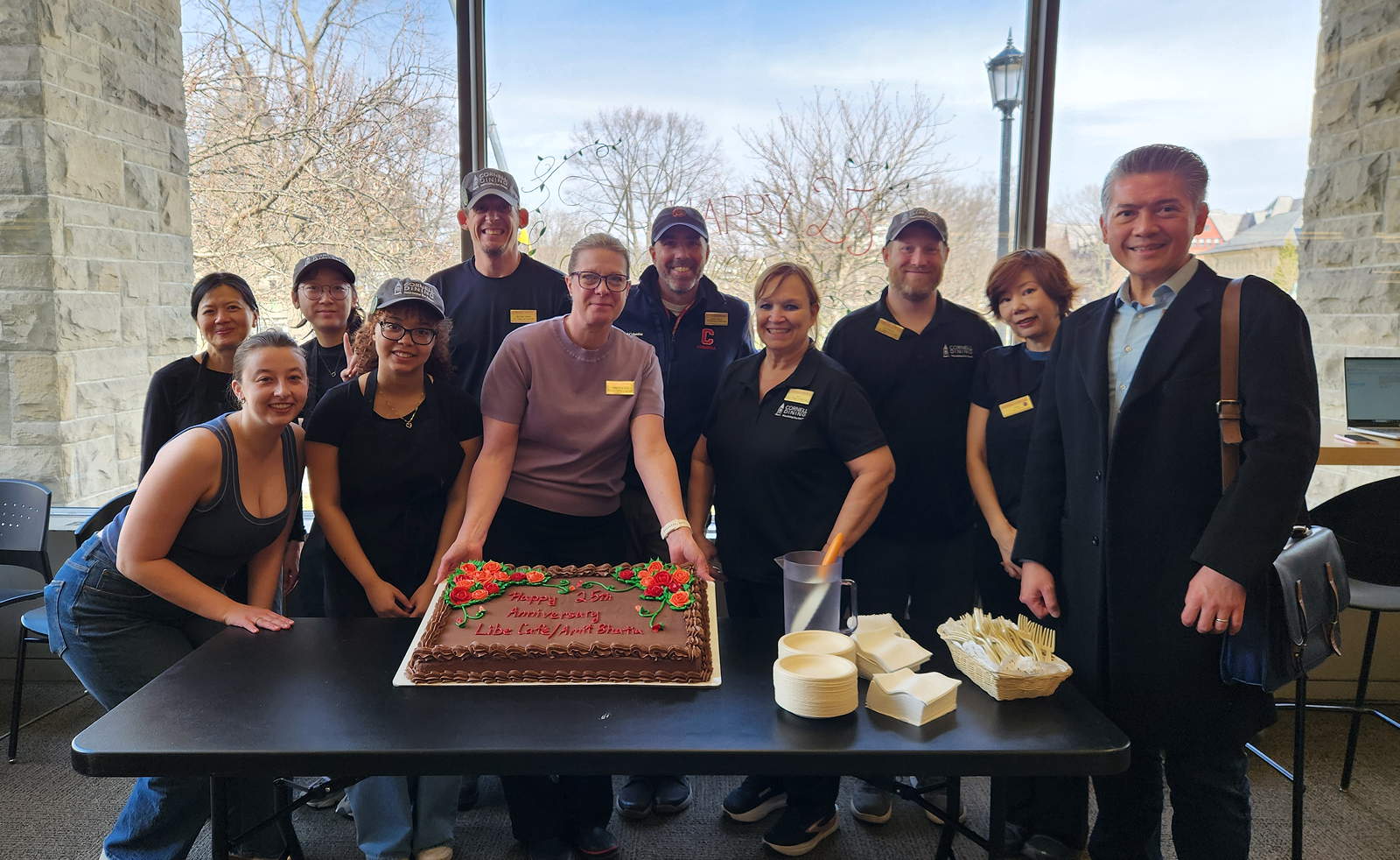 Smiling people wearing cafe uniforms around a table. One person is holding up the edge of a cake.