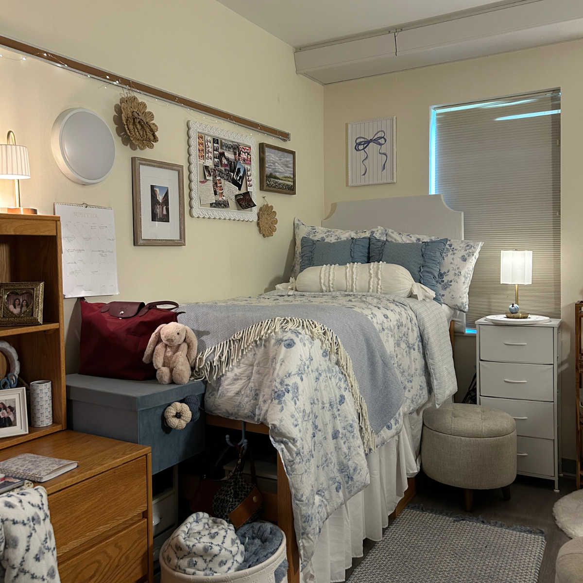 A decorated residence hall room featuring a bed with blue and white floral bedding, a white headboard, and wooden furniture, lit by lamps and string lights