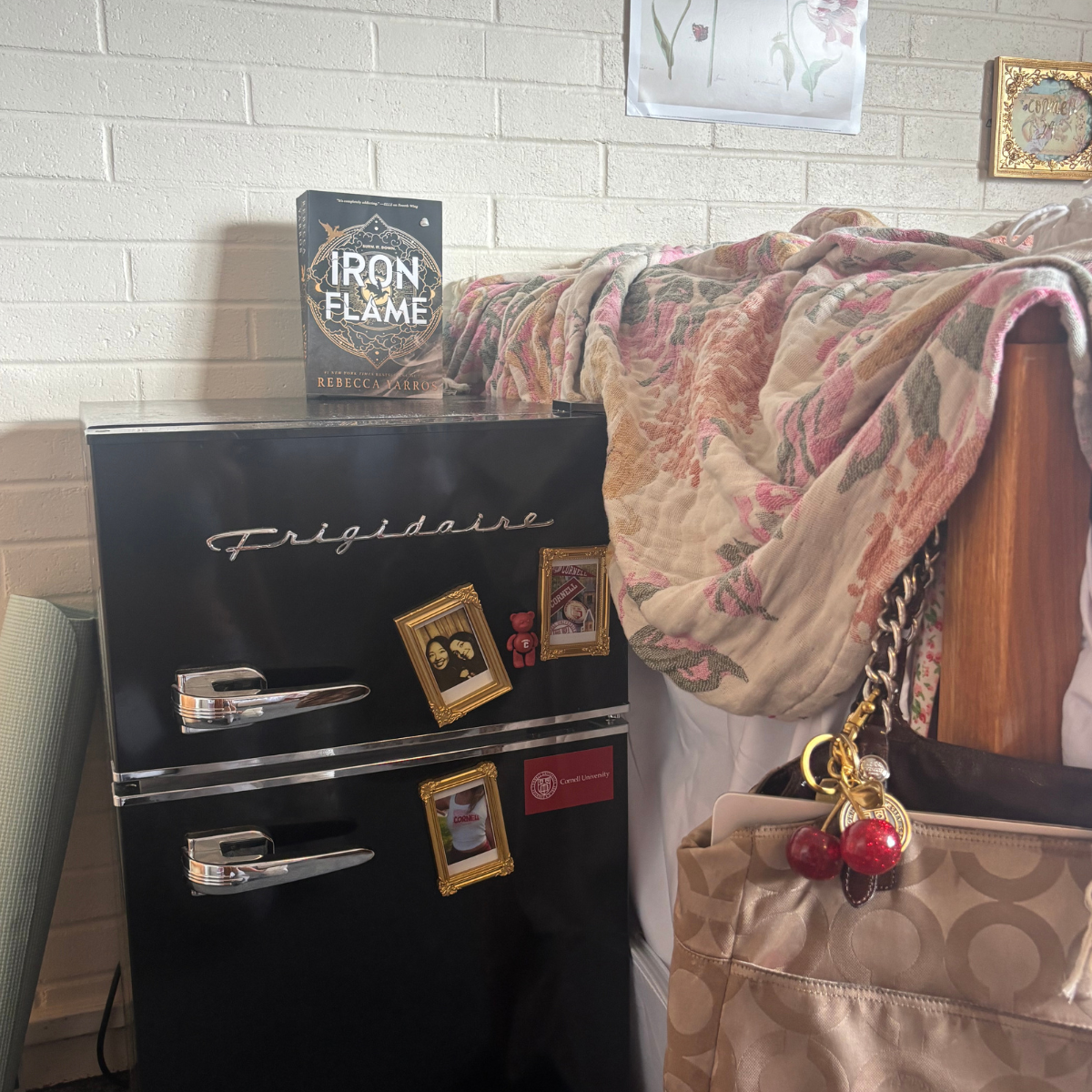 A black Frigidaire mini-fridge next to a bed with a floral quilt, topped with the book "Iron Flame" by Rebecca Yarros