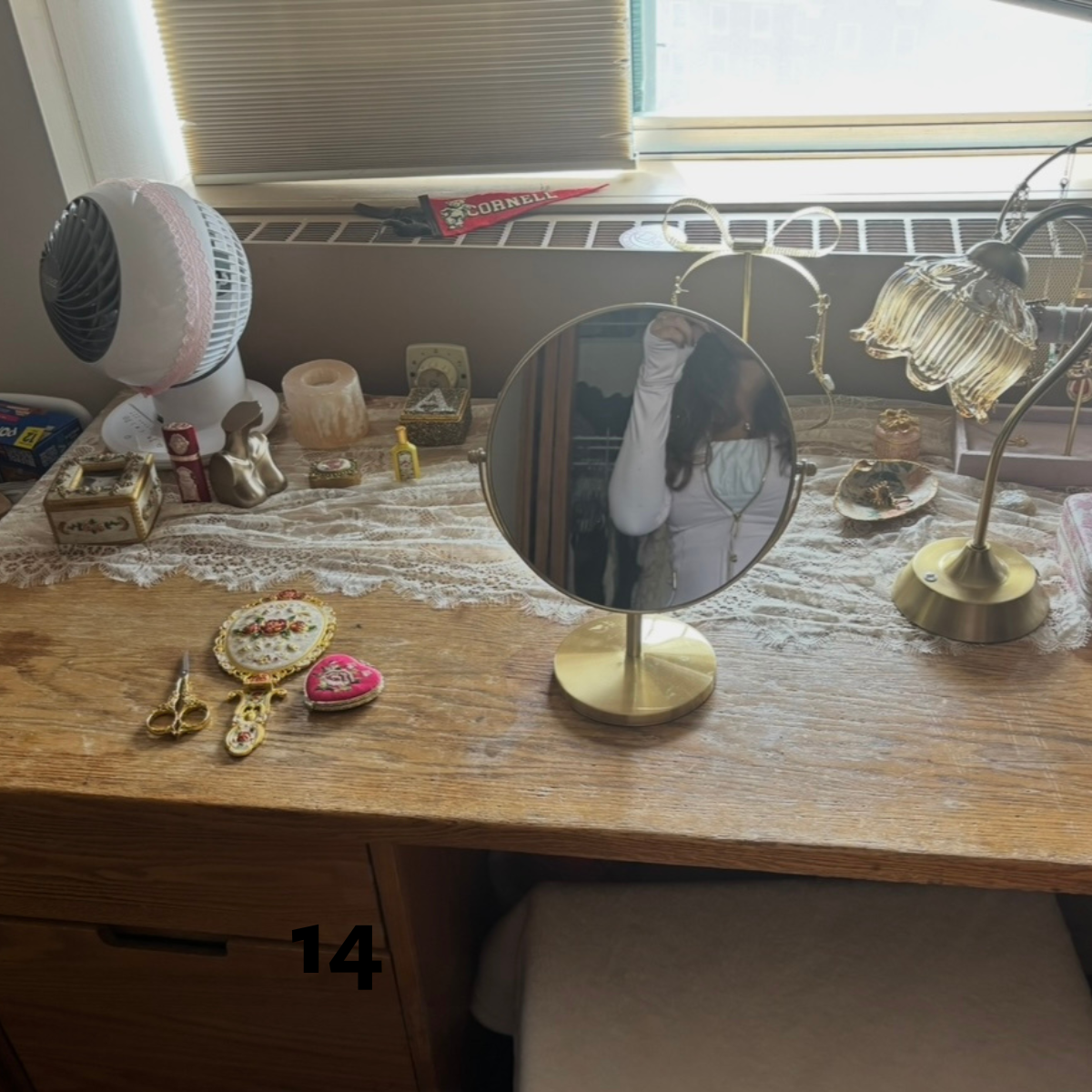 A wooden desk decorated with a gold-framed vanity mirror, a vintage lamp, a small fan, and various small trinkets and jewelry