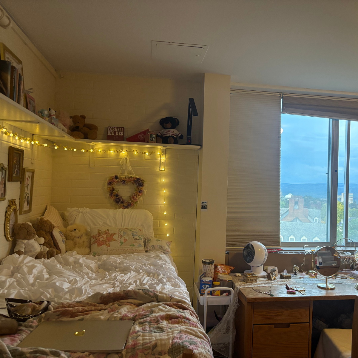 A fully decorated residence hall room featuring a bed with white ruffled bedding and string lights, and a desk next to a large window with a view