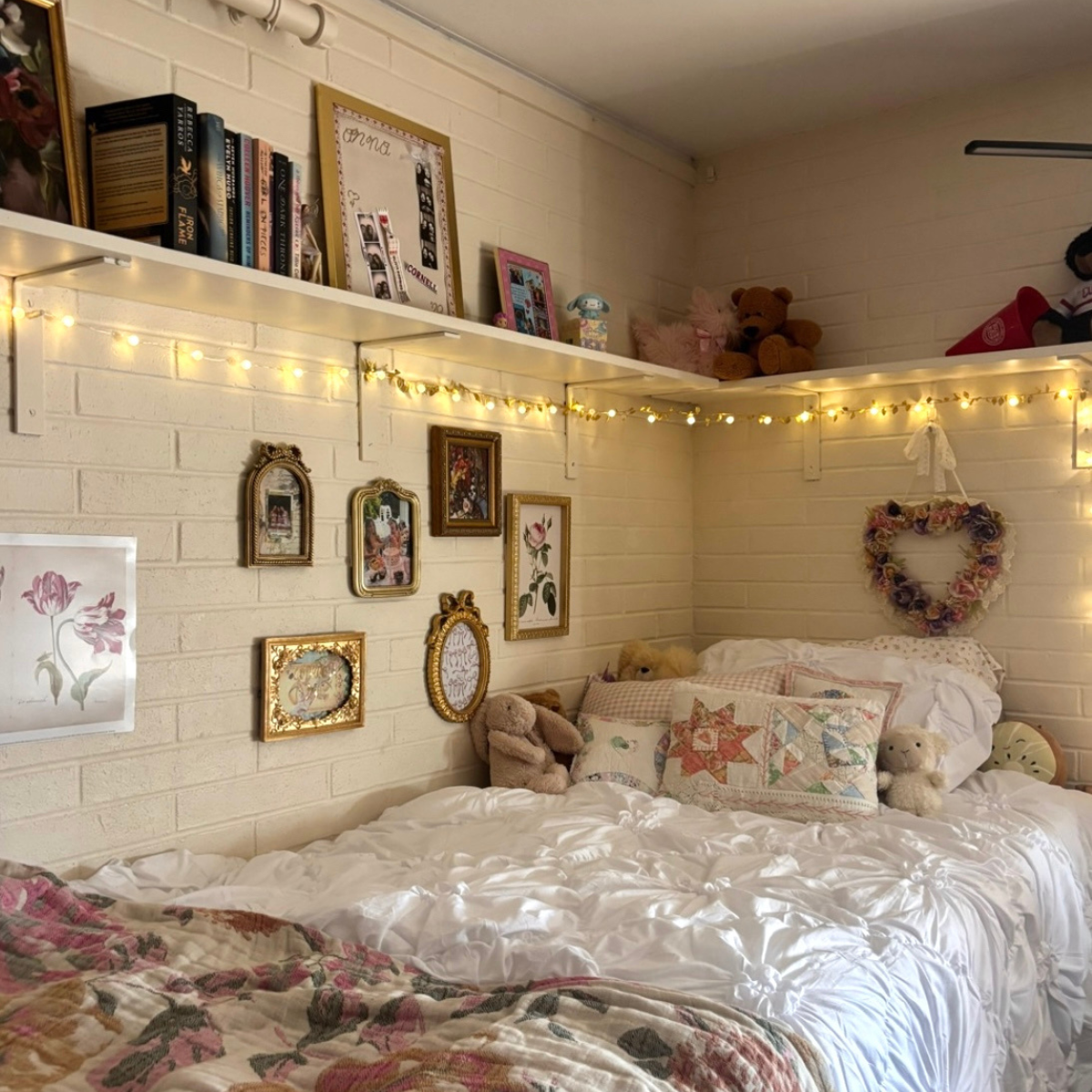 A cozy bed area in a residence hall room with white brick walls, decorated with shelving, gold-framed art, and warm string lights