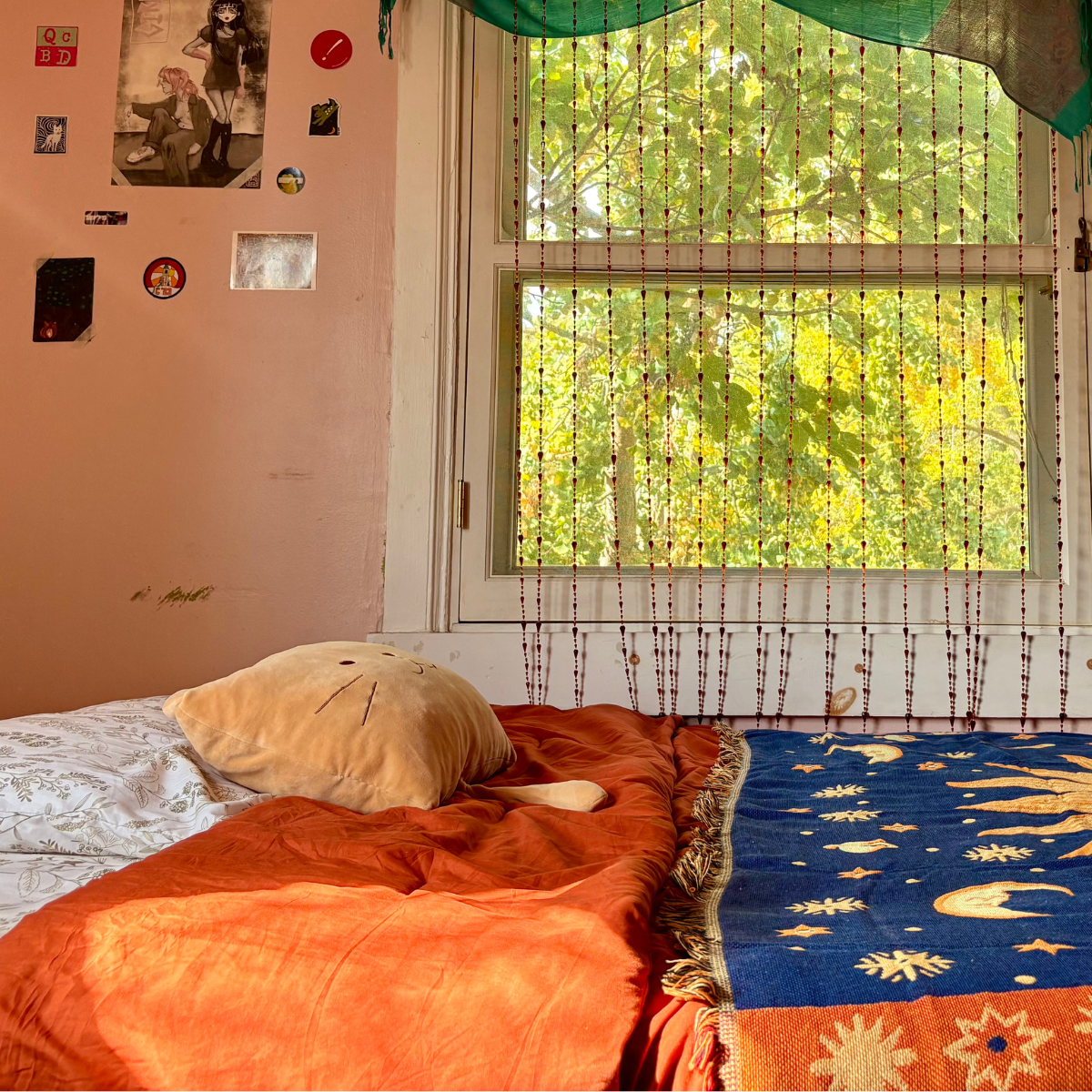 A bed with orange bedding and a celestial-themed throw blanket, next to a window covered by a green beaded curtain with a view of bright green trees