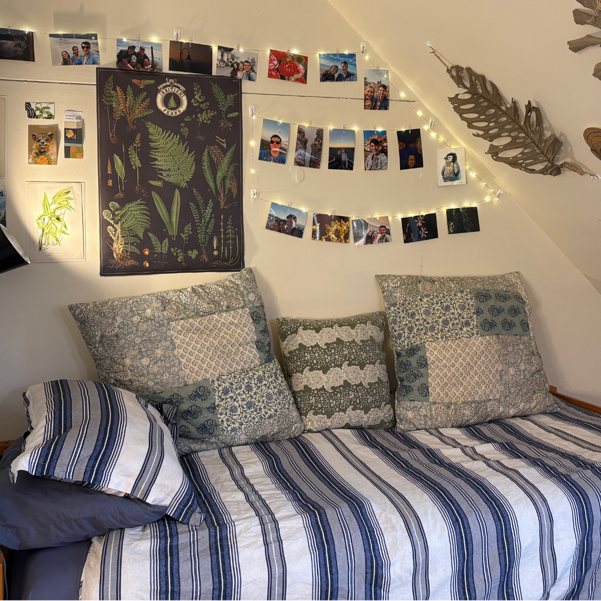 A bed in a room with a sloped ceiling, covered in blue and white striped and patchwork bedding, with a photo display and botanical posters on the wall