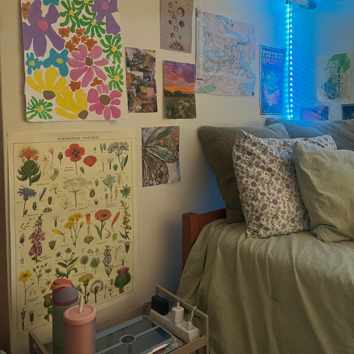 A residence hall room bed with sage green bedding and a large gallery wall featuring colorful, eclectic art, maps, and botanical posters, lit by blue LED lights