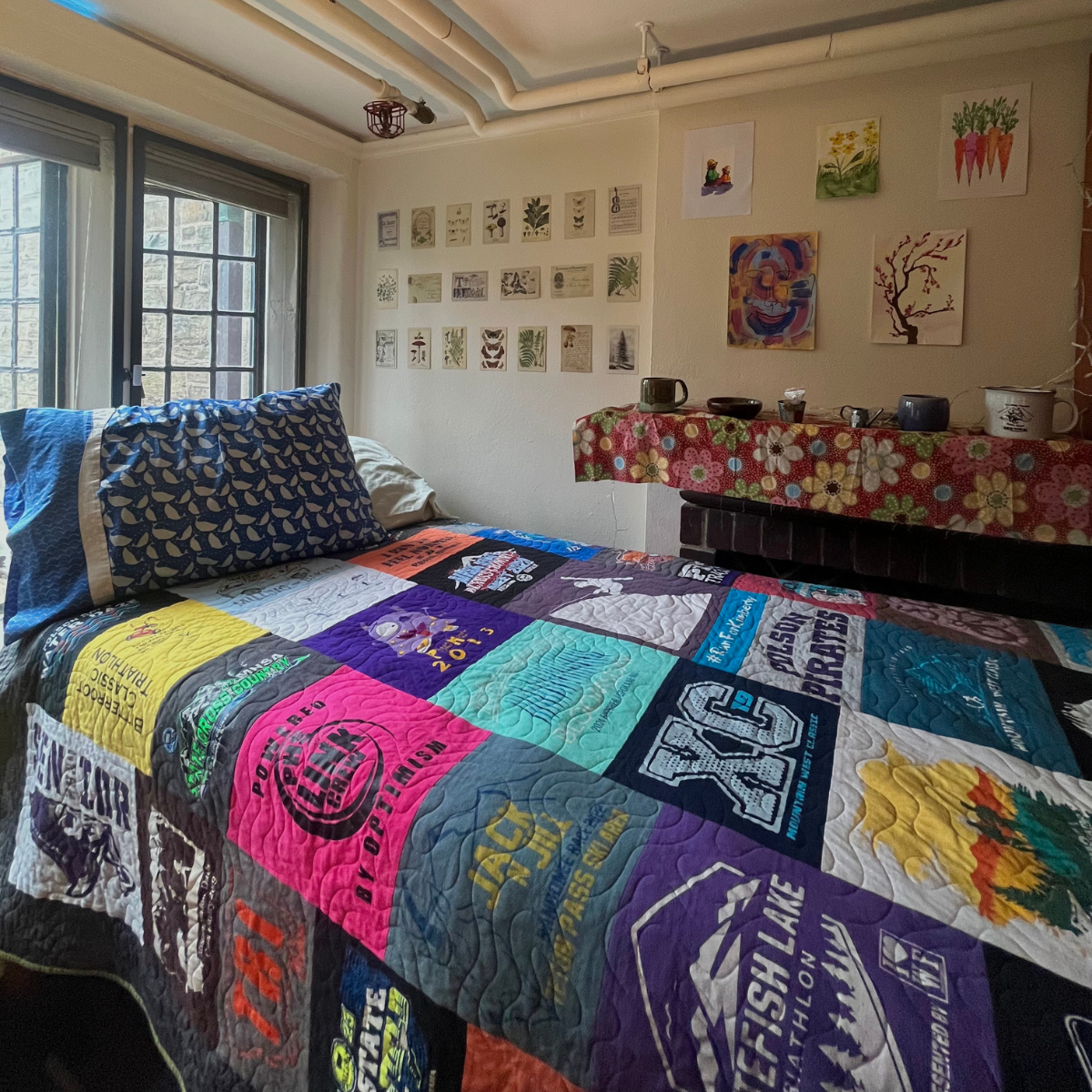 A residence hall room bed covered with a colorful quilt made from various athletic and event T-shirts, next to a large window