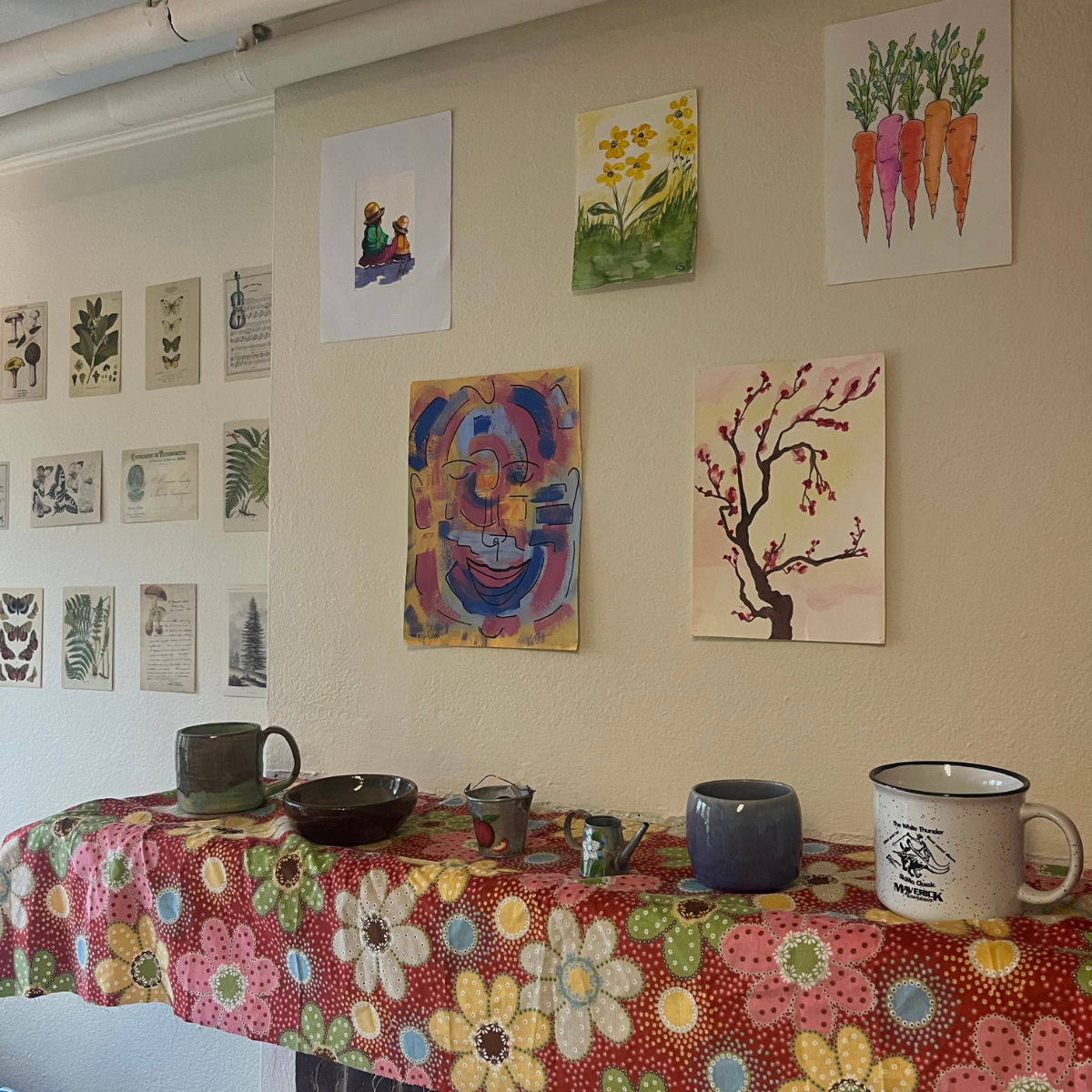 A wall decorated with eclectic watercolor paintings and botanical prints, above a table covered with a floral cloth and ceramic mugs