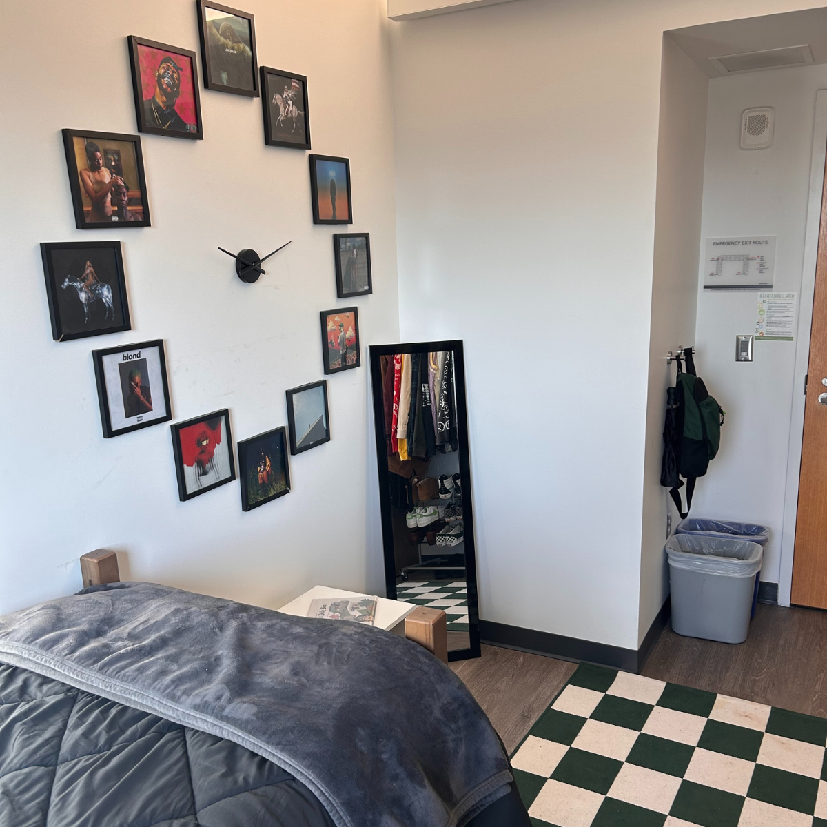 A residence hall room corner with a bed, wall art arranged around a clock, a full-length mirror, and a checkered floor mat near the door