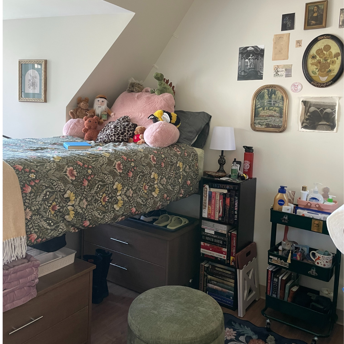 A residence hall room featuring a bed with a floral comforter and stuffed animals, dark wood dresser storage underneath, and a collection of framed art on the wall.
