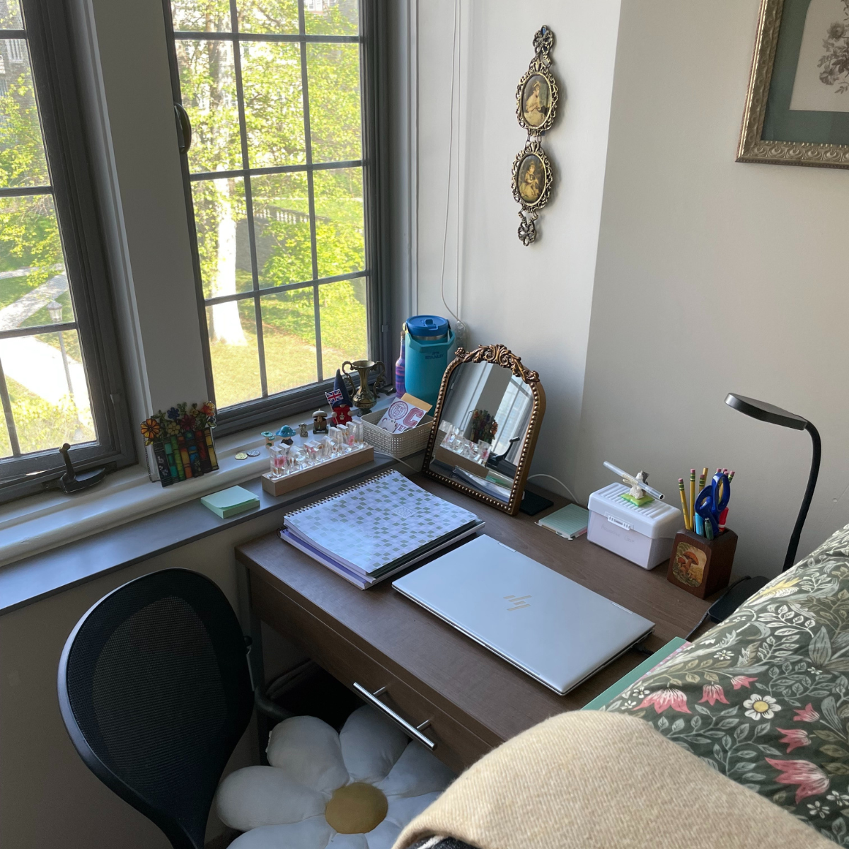 A student desk placed under a large window, featuring a laptop, notebooks, a gold-framed mirror, and various school supplies