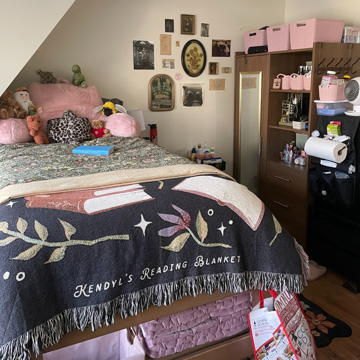 A residence hall room featuring a bed with a dark green patterned blanket and a custom black and tan "Kendyl's Reading Blanket" draped over it. The bed is piled with pillows, including large pink and leopard print cushions. To the right is a tall dark wood wardrobe/storage unit with pink woven storage bins on top and open shelving displaying jewelry and toiletries. A gallery wall of small pictures and framed art hangs above the bed and to the right