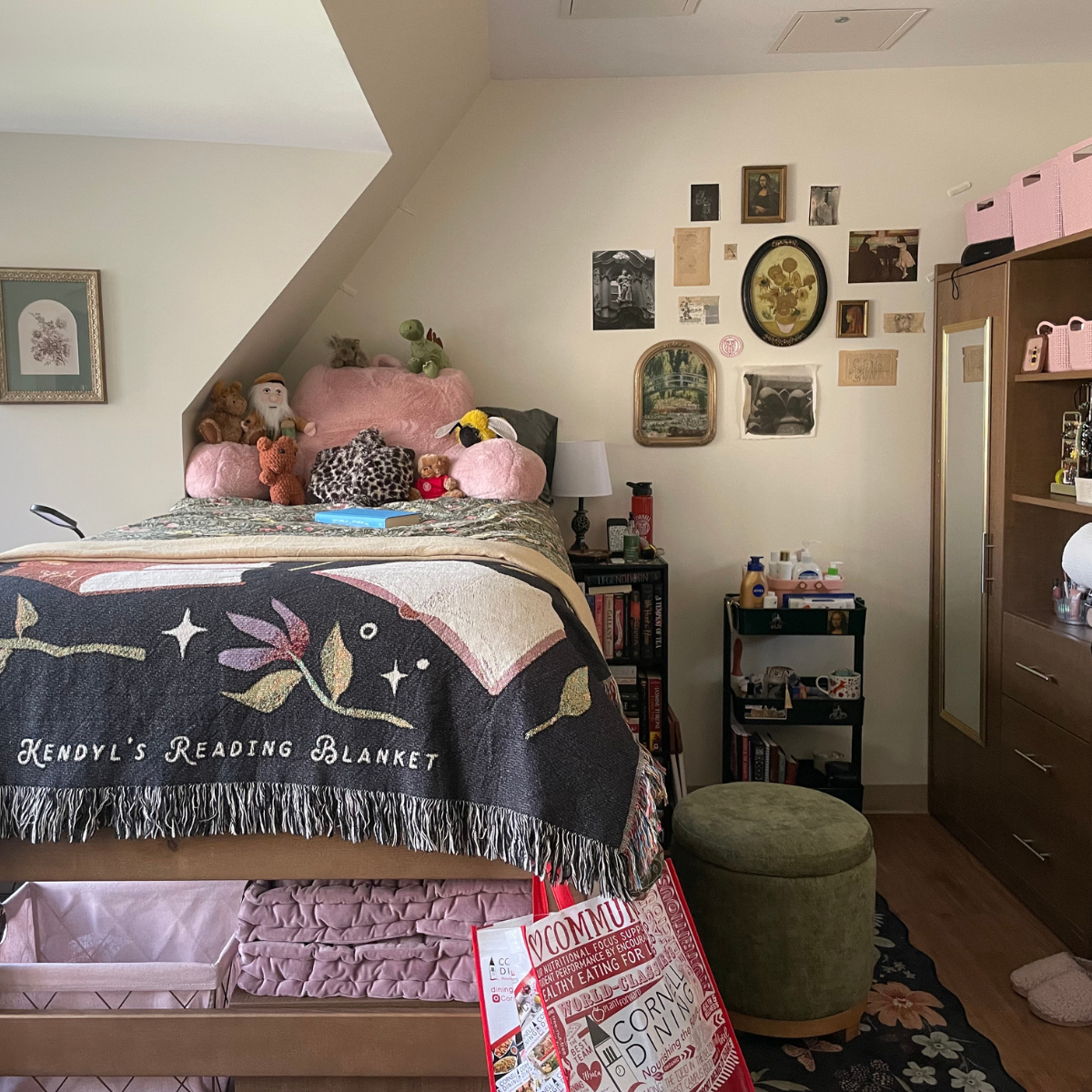 A residence hall room featuring a bed with a dark green patterned blanket and a custom black and tan "Kendyl's Reading Blanket" draped over it. The bed is piled with pillows, including large pink and leopard print cushions. To the right is a tall dark wood wardrobe/storage unit with pink woven storage bins on top and open shelving displaying jewelry and toiletries. A gallery wall of small pictures and framed art hangs above the bed and to the right