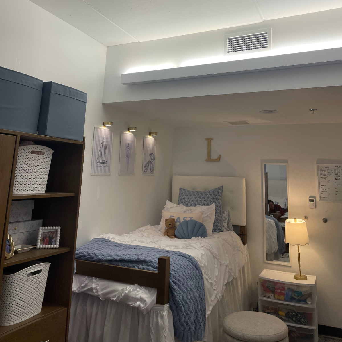 A wide view of a decorated residence hall room with a white bed, navy and blue accents, built-in shelving, and warm lighting