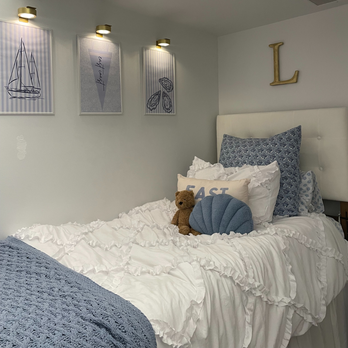A neatly made bed with white ruffled bedding and blue accents, a white tufted headboard, and nautical-themed wall decor