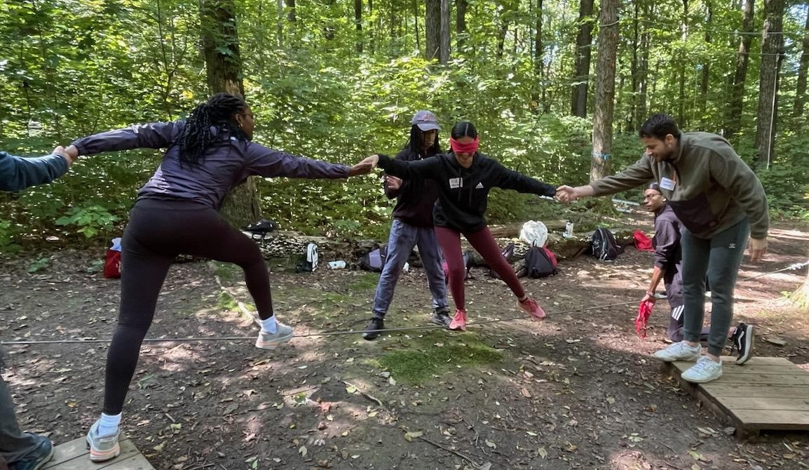 Students outdoors holding hands and balancing on a cable 2 feet above the ground.