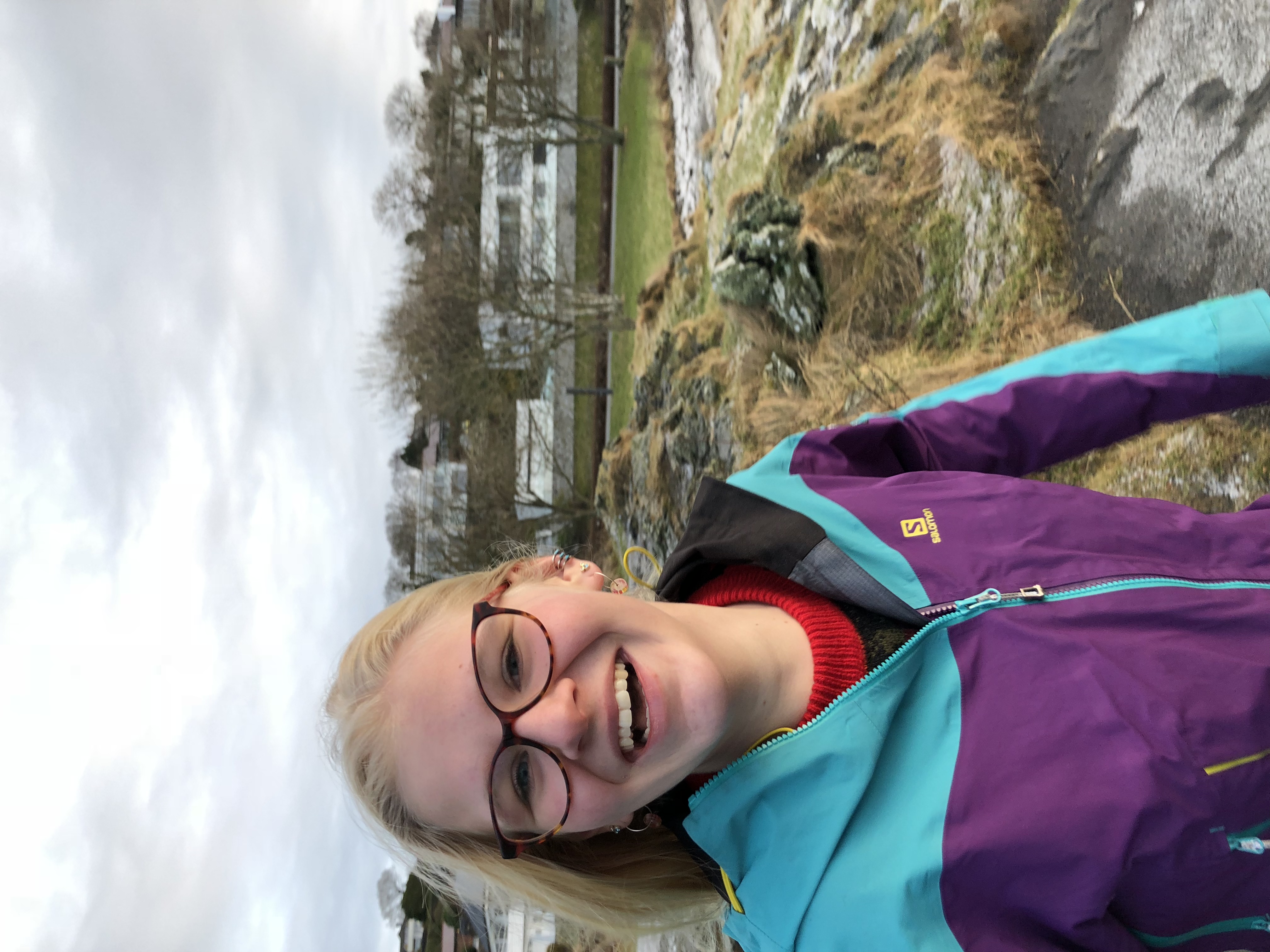 Sarah in brown glasses and a purple and light blue windbreaker standing on a beach in winter with modern white houses in the background