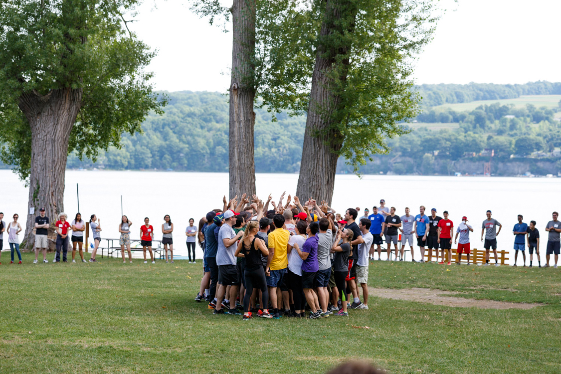 group of 30 people gathered in a tight circle with their hands raised as a large group encircles them near a lake