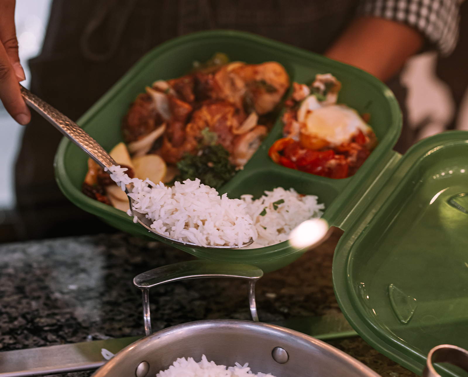 A person uses a serving spoon to put rice into a green plastic takeout container that already has some food in it