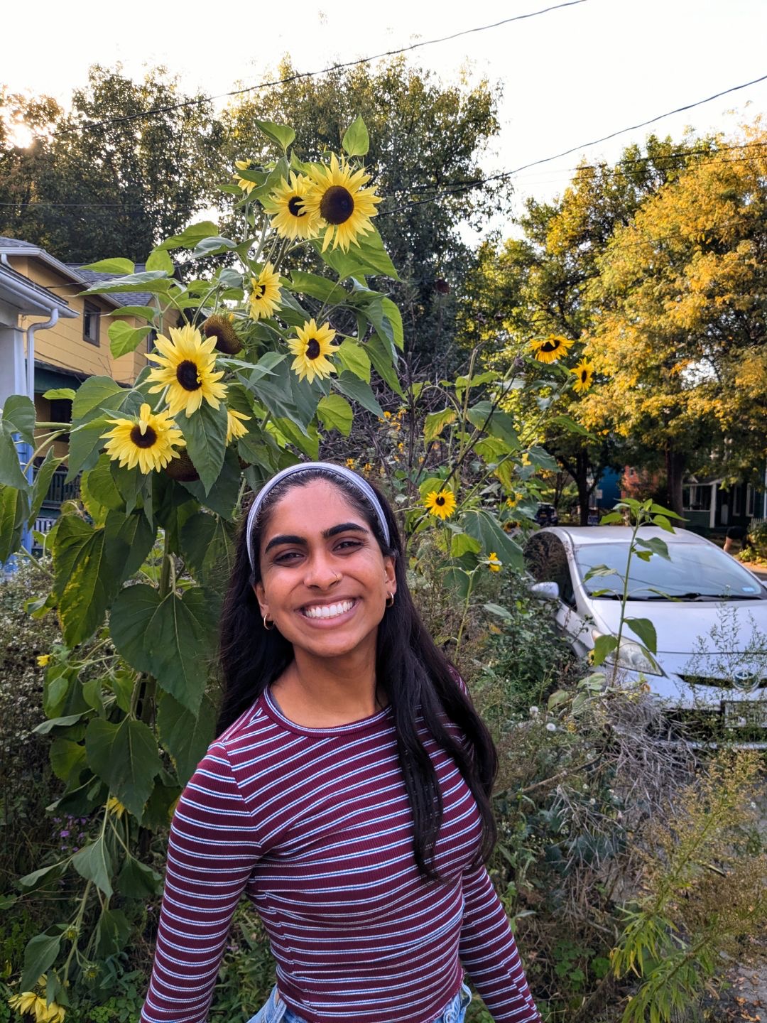 Shivani standing in front of sunflowers wearing a violet headband and striped purple shirt and smiling