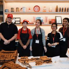 A row of people wearing aprons standing behind a food serving station