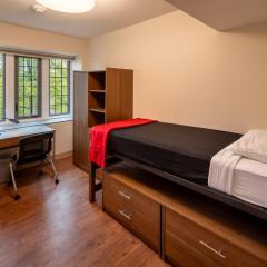 Single room with a bed, desk and chair, drawers, an armoire and a window.