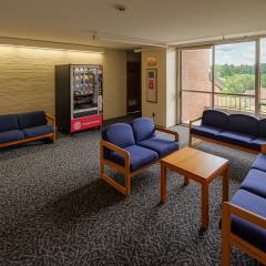 A lounge in Jameson Hall featuring blue lounge chairs, a blue couch, coffee table, drink vending machine, and a large window. 