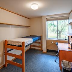 Single with a twin bed, desk, chair, window, and blue carpet.