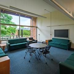 Large lounge with four green couches, a round table with chairs, a tv and a large window over blue carpet.