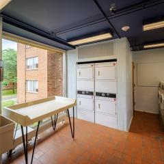 Laundry room with four washers, four dryers, a table to fold laundry in front of a large window, and a sink on red tile floor.