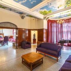A large lounge with a coffee table, two couches, a lounge chair, and a piano in front of a grand window covered in long curtains.