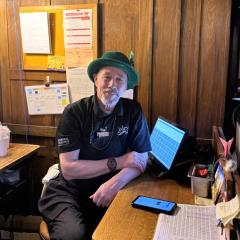 A smiling Garry sits at the door checker station wearing a leprechaun hat