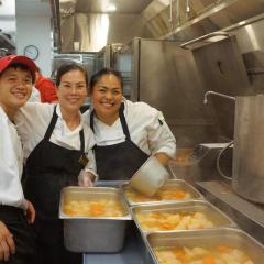 Three people wearing chef coats and aprons in a kitchen with pots of soup