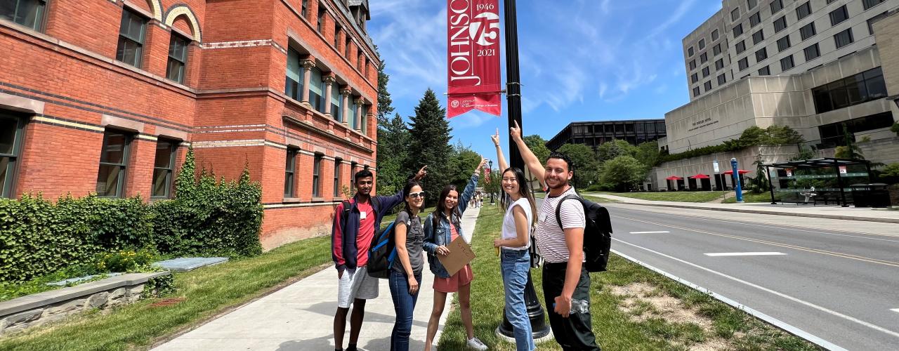 Group of 5 people pointing to Johnson School banner on light post, on Feeney Way outside of Sage Hall at Cornell