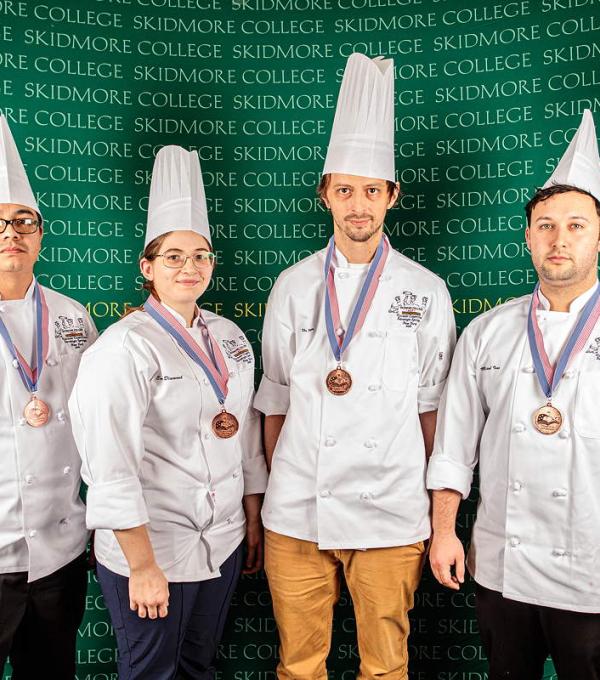 Four people wear chef coats and hats, and medals, in front of a wall that says Skidmore College