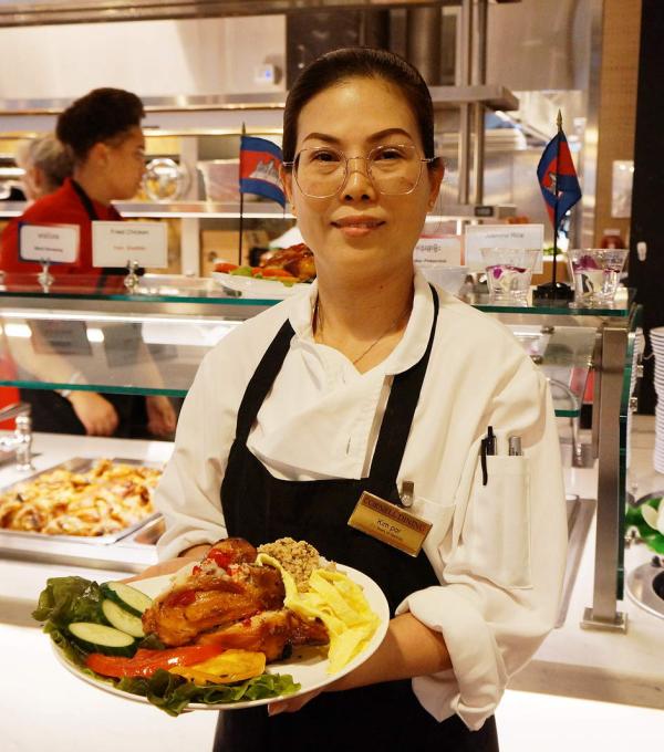 A person in an apron holds a plate of food in front of a serving line