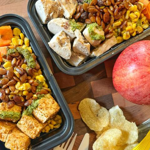 Two takeout containers full of food, with potato chips and an apple on the table next to them