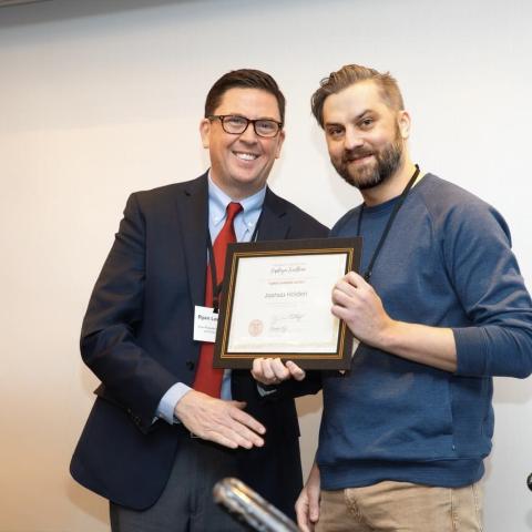 Two people on stage, one holding an award certificate