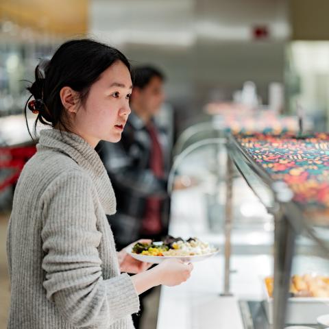 A young woman stands in front of a food serving station with a plate